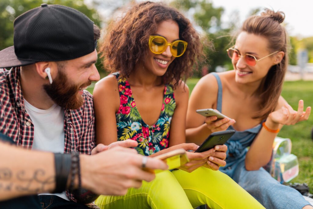 happy young company of smiling friends sitting park using smartphones, man and women having fun together, colorful summer hipster fashion style, communication wireless connecting devices, upswap
