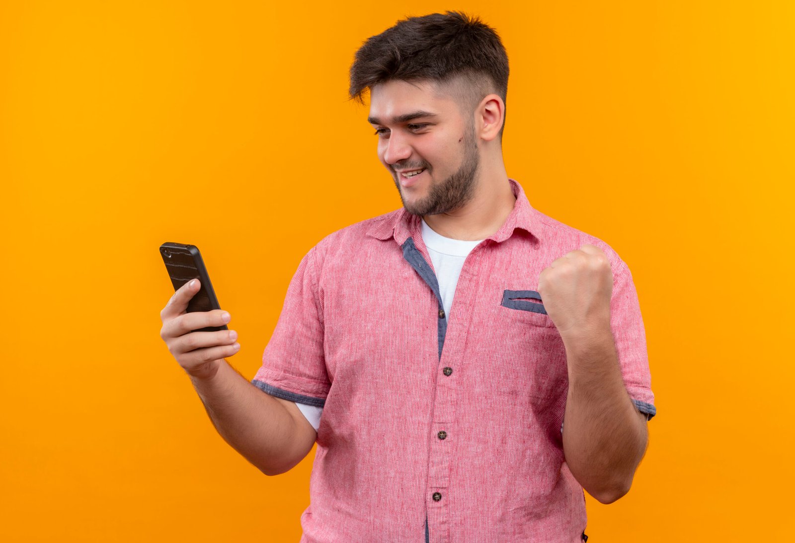 Young handsome guy wearing pink polo shirt looking at phone happy doing did it sign with fist standing over orange wall